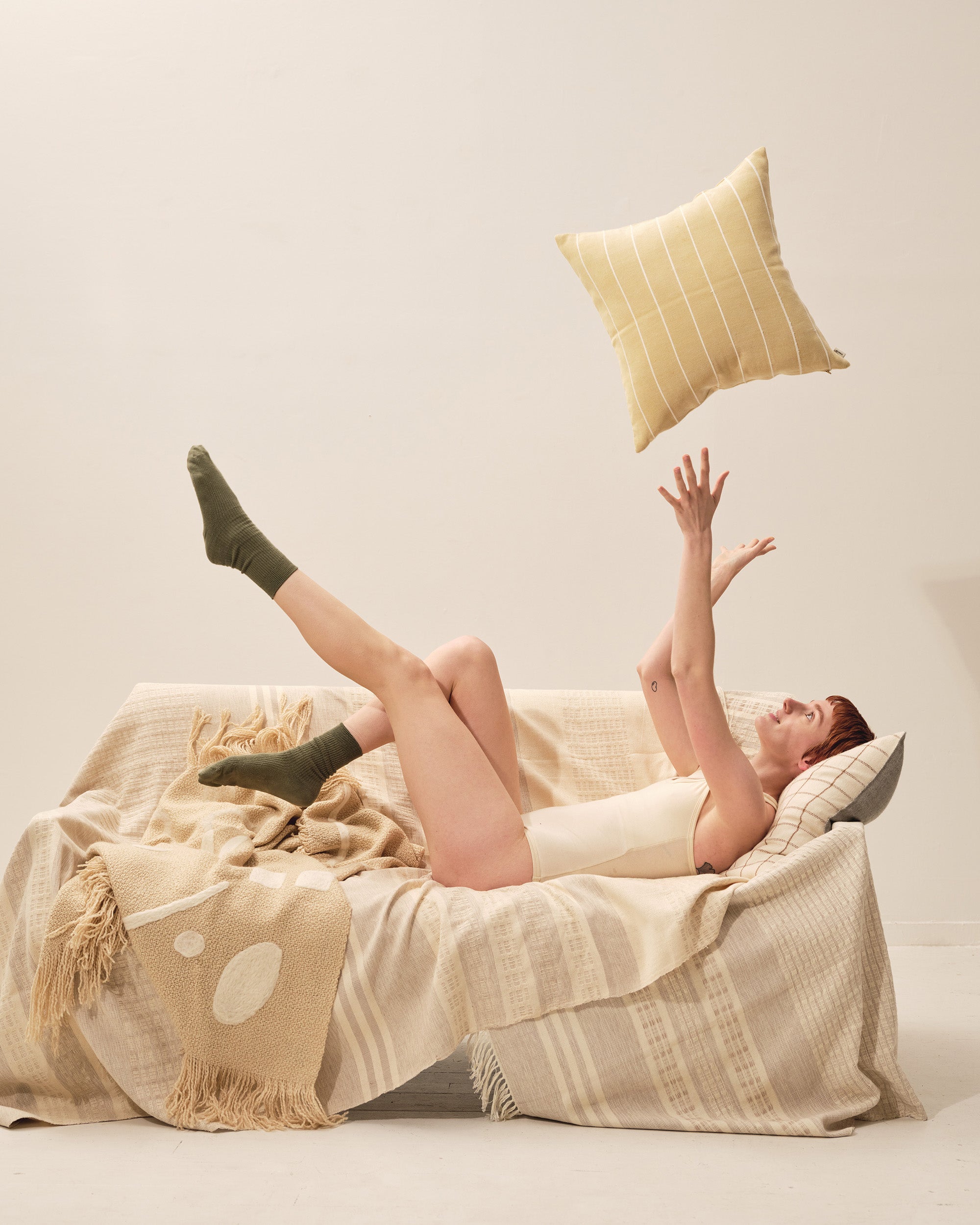 Model on a couch with two neutral ethically handwoven throw blankets tossing a yellow decorative throw pillow in the air. MINNA Abstract Throw Oat